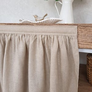 May include: A beige linen curtain panel featuring a gathered top edge with visible stitching. The fabric has a plain weave texture and hangs vertically, covering the front of a wooden surface.