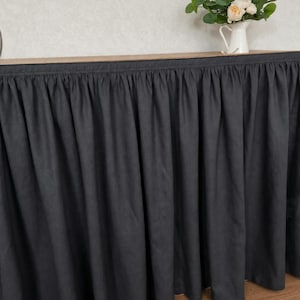 May include: A dark charcoal gray table skirt with a gathered, pleated design attached to a wooden surface. A white ceramic pitcher containing a bouquet of green leaves and light pink flowers sits on the top edge of the wooden table surface.