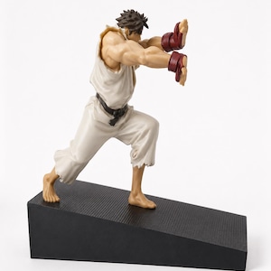 Könnte beinhalten: Eine Figur eines Kampfsportlers in einem weißen Gi mit schwarzem Gürtel und roten Handschuhen, in einer dynamischen Vorwärtsschub-Pose. Die Figur steht auf einem schwarzen, texturierten, keilförmigen Sockel und zeigt detaillierte Muskeln.