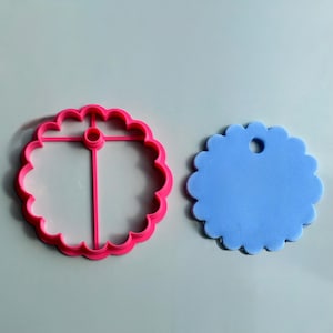Peut inclure: Un emporte-pièce circulaire rose vif avec un bord festonné et une barre transversale centrale dotée d'un petit trou, placé à côté d'une découpe circulaire bleu clair avec un bord festonné et un petit trou près du centre supérieur.