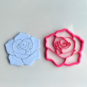 Peut inclure: Une découpe en forme de rose bleu clair faite d'un matériau pressé à côté d'un emporte-pièce rose vif en forme de rose. La découpe présente des lignes de pétales en relief détaillées, tandis que l'emporte-pièce en plastique a un centre creux.