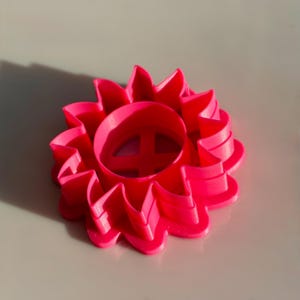 Puede incluir: Cortador de galletas con forma de girasol, impreso en 3D, de color rosa brillante. El cortador presenta un centro circular con una forma de 'A' estilizada y una serie de pétalos curvos. Adecuado para hornear o manualidades.
