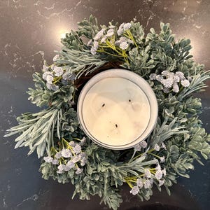 May include: A large, round candle in a metal container, surrounded by a decorative wreath. The wreath is made of green and silver-toned leaves with small white flower accents. The candle has three wicks and a creamy white wax.