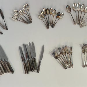 Può includere: Un assortimento di posate antiche, tra cui cucchiai, forchette e coltelli, disposte su una superficie bianca. La collezione presenta varie dimensioni e stili di utensili, mostrando un'estetica vintage. Le posate sono in metallo lucido color argento.