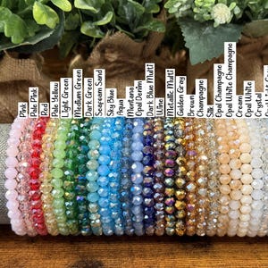 Peut inclure: Une collection de bracelets en perles de cristal &agrave; facettes dispos&eacute;s en rang&eacute;e, chacun &eacute;tiquet&eacute; avec des noms de couleurs comme Pink, Red, Aqua, Wine, Champagne et Hematite. Les bracelets pr&eacute;sentent un spectre de couleurs vari&eacute;es.