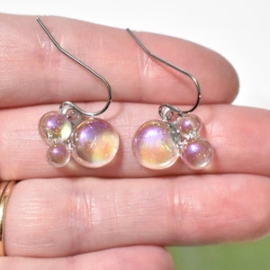 May include: A pair of iridescent bubble earrings. Each earring features a cluster of clear, round beads in varying sizes, with a shimmering, multi-colored finish. The earrings have silver-colored ear wires.
