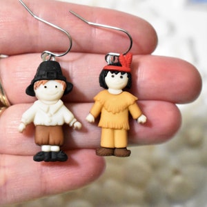 May include: A pair of dangle earrings featuring a pilgrim and an indigenous person. The pilgrim is wearing a black hat, white shirt, brown pants, and black shoes. The indigenous person is wearing a red headband with a feather, a brown shirt with fringe, brown pants, and brown shoes.
