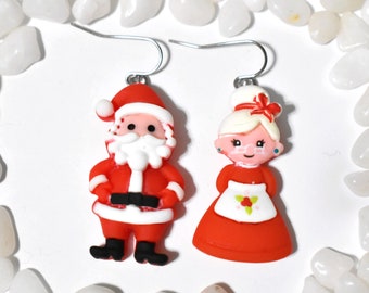 May include: A pair of Christmas-themed earrings featuring Santa Claus and Mrs. Claus. Santa is wearing a red suit with a white trim and a white beard. Mrs. Claus is wearing a red dress with a white apron and a white bow in her hair.