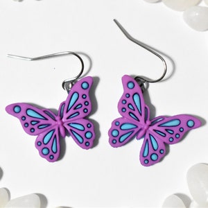 May include: A pair of purple butterfly earrings with blue accents. The earrings have a silver hook.