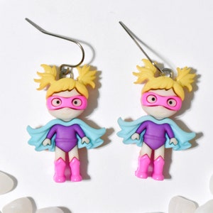 May include: A pair of dangle earrings featuring a cartoon superhero girl with blonde pigtails, a pink mask, a purple leotard, and a light blue cape.