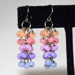 May include: A pair of ombre dangle earrings with peach, pink, lavender, and light blue beads. The earrings are made with silver-toned metal.