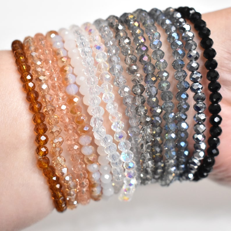 May include: A collection of eleven stretch bracelets featuring faceted crystal beads in a gradient of colors, transitioning from deep amber and peach to translucent white, iridescent clear, various shades of grey, and finally solid black.