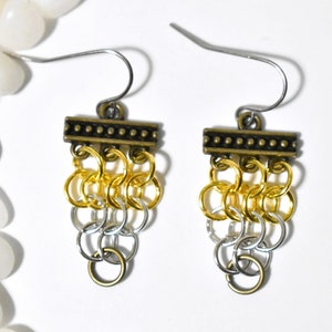 May include: A pair of handmade earrings featuring a cascade of gold and silver-colored chainmail rings. The earrings have a decorative bronze-colored bar at the top with a silver-colored hook.
