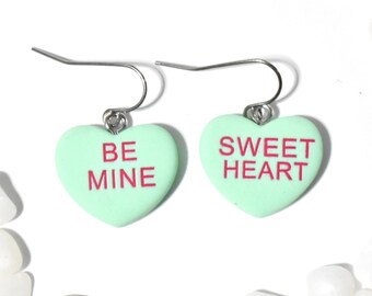 May include: A pair of mint green heart-shaped earrings with silver hooks. The left earring says "BE MINE" and the right earring says "SWEET HEART".