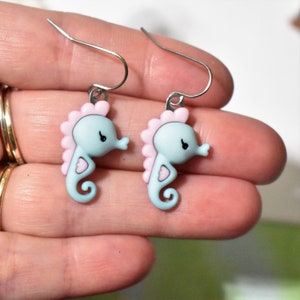 May include: A pair of silver earrings with light blue and pink seahorse charms. The seahorses have a heart-shaped design on their bodies.