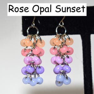 May include: A pair of dangle earrings featuring the text Rose Opal Sunset. The earrings are constructed with silver-toned metal jump rings and multi-colored beads in a gradient pattern of peach, pink, lavender, purple, and light blue.
