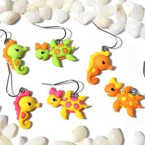 May include: A collection of colorful, handmade earrings featuring seahorse and turtle designs. The earrings are in shades of yellow, orange, pink, and green, with silver-tone ear wires. The earrings are set against a white background with white pebbles.