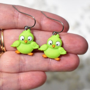 May include: A pair of green bird earrings with orange feet and white eyes. The earrings are made of polymer clay and have silver hooks.