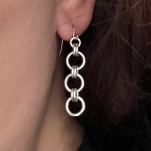 May include: Silver-toned chain earrings. The earrings feature a series of three connected circular links suspended from a hook. The design is simple and elegant, suitable for everyday wear.