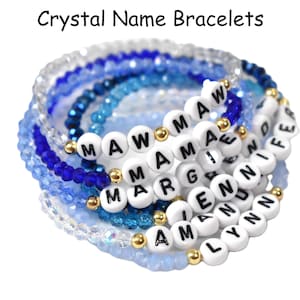 May include: A close-up of a stack of blue and clear crystal bracelets with white beads that spell out names, including "MAW", "MAMA", "MARGIE", "ENN", "AM", and "LYNN".