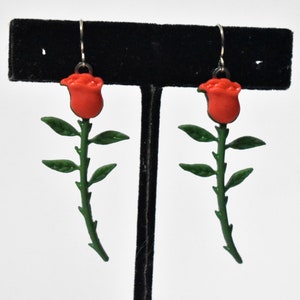 May include: A pair of dangle earrings featuring a red rose with green leaves and stem. The earrings are made of metal and have a silver finish.