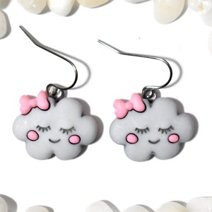 May include: A pair of cloud-shaped earrings. Each earring features a gray cloud with a pink bow, closed eyes, and pink cheeks. The earrings have silver-colored fish hook ear wires.
