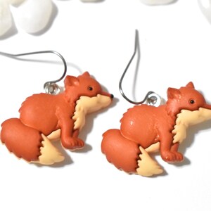 May include: A pair of fox earrings. Each earring features a small, orange fox with a cream-colored chest and belly. The fox is in a sitting position with a curved silver earring hook.