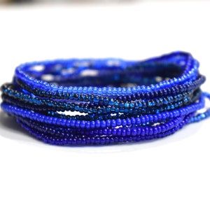 May include: A multi-strand bracelet composed of numerous small, round glass beads in various shades of blue, ranging from light sky blue to deep navy. The beads are strung together in a circular, layered formation, creating a dense, textured appearance.