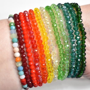 May include: A collection of eleven beaded bracelets featuring faceted glass beads in a spectrum of colors including red, orange, yellow, light green, dark green, and a multi-colored strand with white, blue, and peach tones arranged in a row.