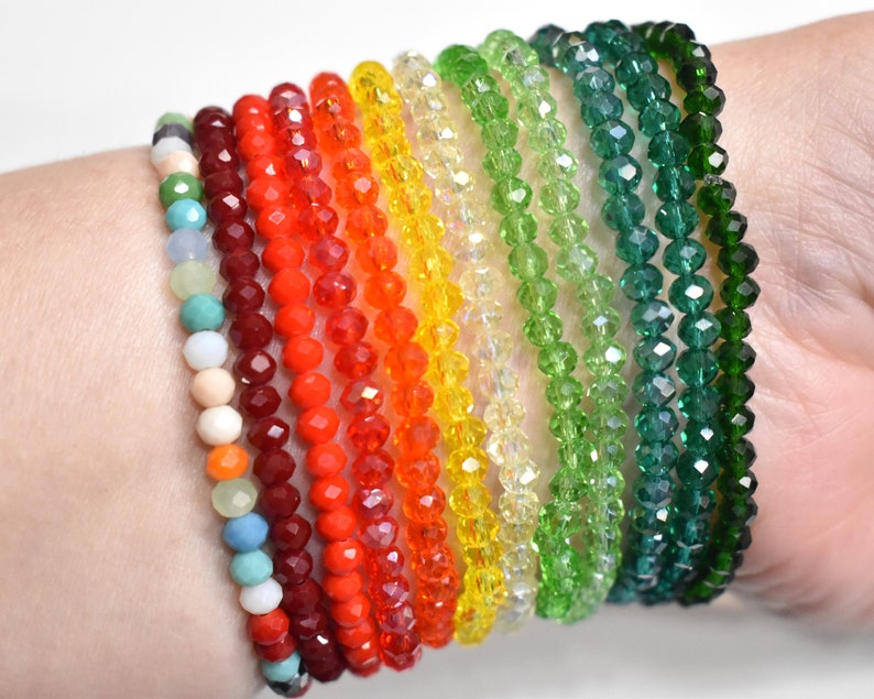 May include: A collection of eleven beaded bracelets featuring faceted glass beads in a spectrum of colors including red, orange, yellow, light green, dark green, and a multi-colored strand with white, blue, and peach tones arranged in a row.