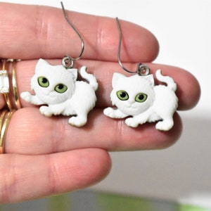 May include: A pair of white kitten dangle earrings with green eyes. The earrings are made of plastic and have a silver hook.