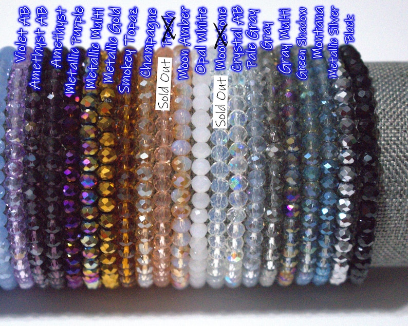 May include: A collection of faceted crystal bead bracelets arranged in a row, featuring various colors including violet, amethyst, metallic purple, gold, champagne, amber, white, gray, green, and black, with text labels identifying each color variant.