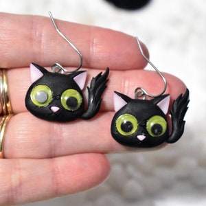 May include: A pair of black cat earrings with googly eyes and pink ears. The earrings are made of polymer clay and have silver hooks.