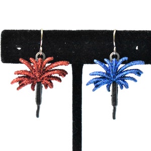 May include: A pair of dangle earrings featuring a red and blue glitter firework design. The earrings are on a black display stand.
