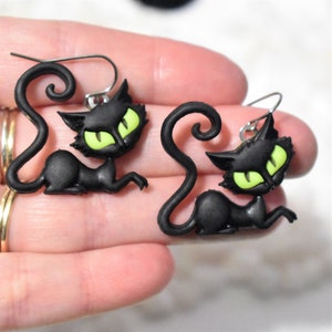 May include: A pair of black cat earrings with bright green eyes. The cat's tail is curled, and the earrings have silver-colored hooks. The earrings are a fun accessory for Halloween or everyday wear.