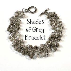 May include: A bracelet with a toggle clasp, featuring clusters of gray and white beads. The beads are strung on silver-colored chain links. The text "Shades of Grey Bracelet" is displayed.