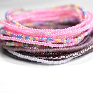 May include: A stack of colorful beaded bracelets in shades of pink, purple, and gray. The bracelets are made of small, round beads and are arranged in a layered fashion.