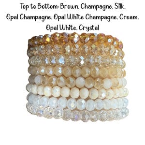 Peut inclure: Une pile de huit bracelets de perles avec des perles en cristal facett&eacute;es dans un d&eacute;grad&eacute; de couleurs. Le texte indique : Top to Bottom: Brown. Champagne. Silk. Opal Champagne. Opal White Champagne. Cream. Opal White. Crystal. Chaque bracelet est compos&eacute; de perles rondes et r&eacute;fl&eacute;chissantes.