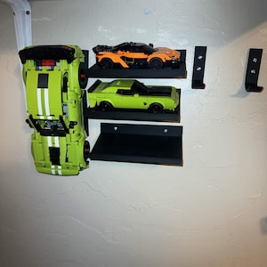 May include: Three toy cars, in green, orange, and yellow, are displayed on black shelves mounted on a wall. The green car is positioned vertically. The orange car is on the top shelf, and the yellow car is on the middle shelf.