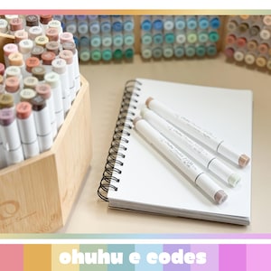 May include: A selection of Ohuhu markers in a wooden organiser, alongside a spiral-bound notebook and several markers on top. The markers are white with coloured tips, and the organiser is filled with various shades. The text "ohuhu e codes" is at the bottom.