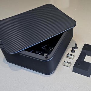 May include: A black rectangular box with a carbon fibre textured lid, partially open. The box has internal supports. Several small metal and black components are arranged to the right of the box.