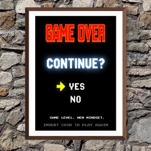 May include: A framed print with a black background and pixelated white text that reads "GAME OVER CONTINUE?" with "YES" and "NO" options. The text "SAME LEVEL. NEW MINDSET. INSERT COIN TO PLAY AGAIN" is also present. The frame is brown.