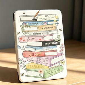May include: A white e-reader case featuring a colorful illustration of a stack of books. The spines are labeled with titles including Midnights, evermore, folklore, Lover, reputation, 1989, RED, Speak Now, FEARLESS, and Taylor Swift, with floral accents.