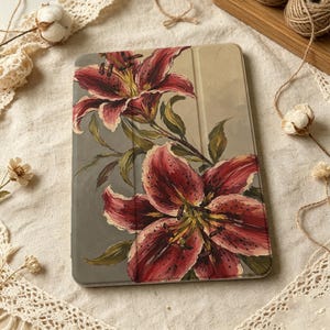Puede incluir: Una funda rectangular para tableta con un diseño pintado de dos grandes lirios de color rojo intenso con motas oscuras y hojas verdes sobre un fondo gris y beige apagado. La funda tiene una superficie lisa con una línea de pliegue vertical.