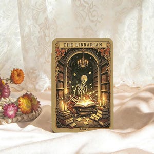 Könnte beinhalten: Eine rechteckige Tarotkarte mit dem Titel THE LIBRARIAN und einer zentralen Illustration eines Skeletts, das in einer Bibliothek sitzt und ein leuchtendes Buch liest. Die Szene zeigt Bücherregale, einen Kronleuchter und Kerzen in Gold, Braun und Schwarz.