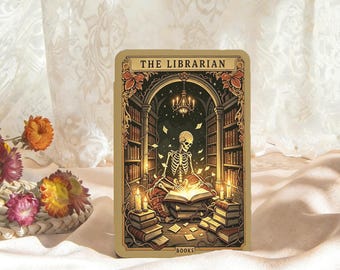 May include: A rectangular tarot card titled THE LIBRARIAN with a central illustration of a skeleton sitting in a library reading a glowing book. The scene features bookshelves, a chandelier, and lit candles, rendered in shades of gold, brown, and black.