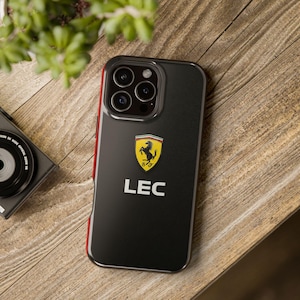 May include: Black phone case with a red trim, displaying the Ferrari logo and the name "LEC" in white. The case is set on a wooden surface, with a camera and foliage visible.