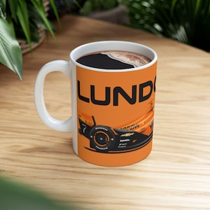 May include: White ceramic coffee mug with the word "LUND" in black block letters on an orange background. The mug features a race car design with the number 7. The mug is filled with dark coffee.