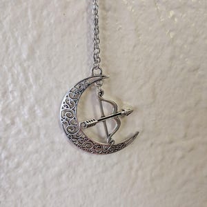 May include: Silver-toned pendant necklace featuring a crescent moon with intricate scrollwork and a bow and arrow design. The pendant hangs from a delicate chain, creating a celestial-themed accessory. The necklace is displayed against a neutral background.