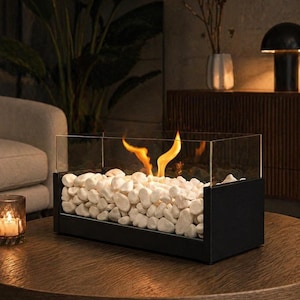 May include: A rectangular tabletop fireplace featuring a black metal base and clear glass panels. The interior is filled with smooth white decorative stones, and a small, bright orange flame rises from the centre of the stone bed.